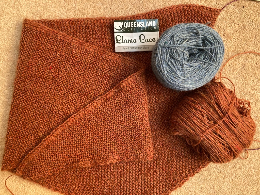 A garter stitch shawl in a copper colour lies draped on the carpet. The ball band is on top, next to a silvery-blue ball of the same yarn which will be used for the lace border.