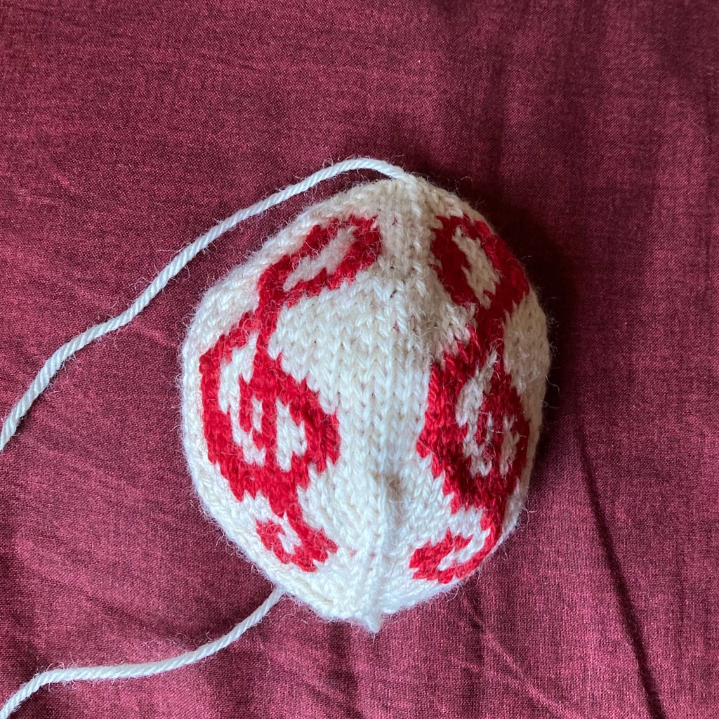 A hand knitted stranded Christmas bauble lies on maroon fabric. The background is cream with two bright red treble clefs showing. The bauble has not yet been stuffed and finished.