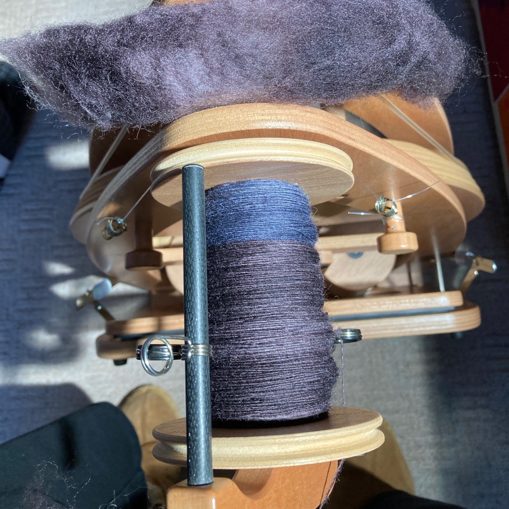 An overhead view of a spinning wheel with a half-filled bobbin of singles. The yarn is two shades of purple, with a more bluey shade in the top quarter of the bobbin. A chunk of grey-purple fibre rests on top of the wheel, on the band.